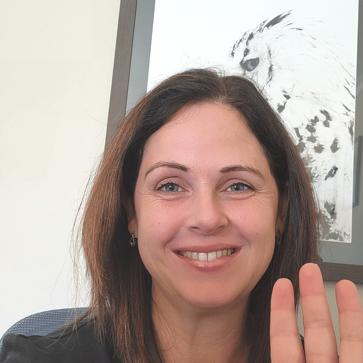 Picture of female small business copywriter waving and smiling at the camera. She has dark hair and is seated in front of a black and white picture of an owl